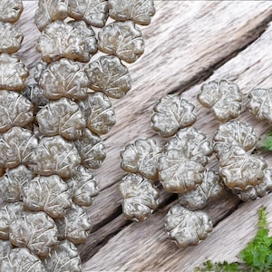May include: Close-up of multiple silver-colored leaf-shaped beads. The beads are arranged on a weathered wooden surface, with some beads clustered together and others scattered. Green foliage is visible in the background.