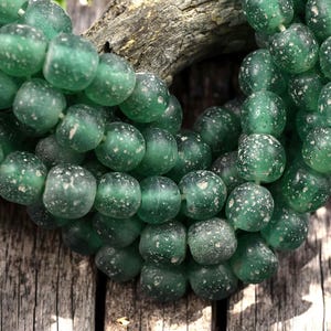 May include: A strand of green glass beads with white speckles. The beads are round and have a slightly irregular shape.