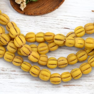 Puede incluir: Primer plano de cuentas amarillas con rayas marrones oscuras. Las cuentas tienen forma ovalada y están ensartadas, creando un accesorio texturizado. Las cuentas están sobre una superficie blanca.