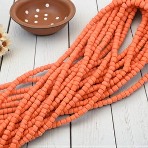 May include: Strands of coral-colored beads are displayed on a white wooden surface. A small, brown bowl with white dots and a small bouquet of white and yellow flowers are also present. The beads appear to be made of a matte material.