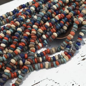 May include: A collection of small, round, disc-shaped beads in a variety of colors including terracotta orange, deep blue, light turquoise, and speckled cream. The beads have a porous, textured surface and are strung together in multiple strands.
