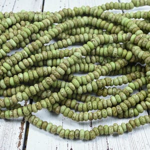 May include: Strands of olive green beads with a speckled, aged appearance. The beads are small, round, and strung together, creating a textured, organic look. The beads are displayed on a white wooden surface.