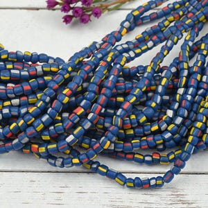 May include: Strands of blue, red, yellow, and white beads. The beads are rectangular and strung together, creating a textured and colorful pattern. The beads are arranged on a white wooden surface.