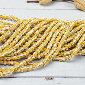 May include: Strands of yellow, white, and green glass beads. The beads are barrel-shaped and strung together, creating a textured and colorful display. The beads are arranged on a white wooden surface.