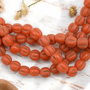 May include: Strands of orange-red beads with a pumpkin-like shape and carved lines. The beads are strung together, creating a necklace or bracelet. The beads are on a white wooden surface.