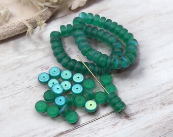 May include: An assortment of green glass beads, including strands of round beads and individual disc-shaped beads. A gold-colored beading needle is also present. The beads have a translucent quality, with hints of blue and green hues.