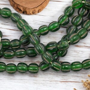 May include: Strands of green glass beads with a ribbed design. The beads are strung together on a white string. The beads are oval-shaped and have a dark green color with lighter green and tan stripes. The beads are displayed on a white wooden surface.