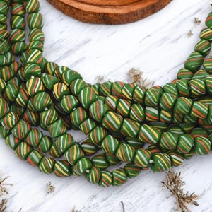 May include: Strands of green, barrel-shaped beads with colorful stripes of white, yellow, and orange. The beads are arranged on a white wooden surface, with a wooden coaster in the background. These beads are ideal for jewelry making.