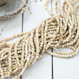 May include: A large collection of small, round beads strung together on multiple strands. The beads feature a variegated color palette, alternating between shades of off-white, cream, and light tan, creating a textured, earthy appearance.