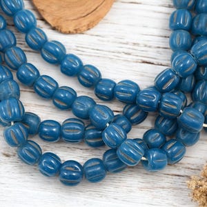 Puede incluir: Primer plano de varios hilos de cuentas de vidrio azules. Cada cuenta tiene forma redondeada con ranuras verticales y un color ligeramente más claro en las ranuras. Las cuentas están ensartadas en un hilo blanco, sobre una superficie de madera clara.