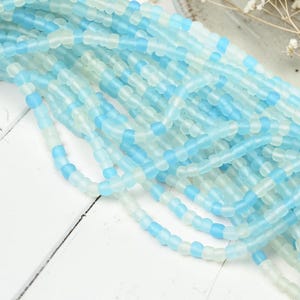 May include: A collection of multiple strands of small, round beads in alternating shades of opaque sky blue and translucent frosted white. The beads are strung together in long, continuous lines, creating a textured, layered appearance.