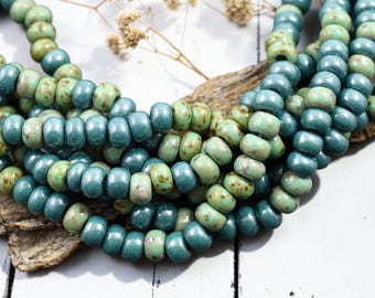 May include: Strands of teal and olive green beads. The beads are cylindrical with a slightly irregular shape, giving them a rustic appearance. The beads are arranged in multiple strands, creating a layered effect. The background is a mix of wood and white.