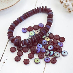 May include: A dark purple beaded bracelet and loose beads. The beads are a flat, disc shape with a hole in the center. Some beads have an iridescent sheen, reflecting colors like blue and green. The bracelet is strung together, while the other beads are scattered.