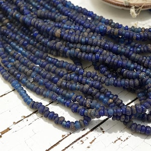 May include: A collection of multiple strands of small, round, disc-shaped beads in varying shades of deep blue. The beads feature a textured, weathered surface with some translucent blue sections and some opaque, dark, matte blue segments.