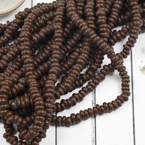 May include: Close-up of several strands of dark brown wooden beads. The beads are small, round, and strung together, creating a textured and organic appearance. The beads are scattered on a white surface, suggesting a craft or jewelry-making theme.
