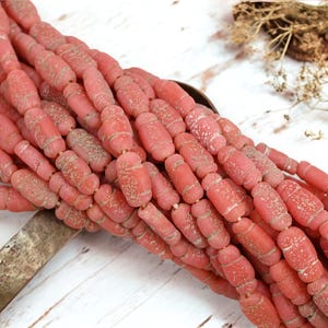 May include: Strands of pink, barrel-shaped beads with a textured, aged appearance. The beads are strung together, creating a dense, tactile display. The color is a muted, dusty rose, with subtle variations in tone. The beads are arranged on a white wooden surface.