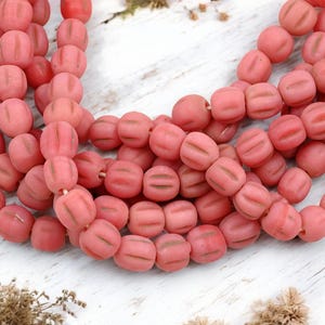 May include: Strands of pink beads with a matte finish. Each bead is oval-shaped with three horizontal brown lines. The beads are strung together, creating a necklace or bracelet.