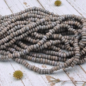 May include: Strands of gray and tan-colored beads with a textured, speckled appearance. The beads are round and uniform in size, creating a consistent pattern. The beads are arranged on a white wooden surface.