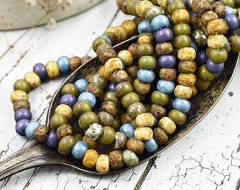 May include: A collection of round, multi-colored beads in shades of olive green, muted blue, purple, and speckled tan. The beads are strung together and piled inside a tarnished, vintage-style metal spoon with a dark, weathered finish.