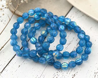 May include: A set of three stretch bracelets composed of translucent blue, ribbed, spherical beads. Each bead features a subtle iridescent finish that catches the light, creating a shimmering effect across the circular, linked jewelry pieces.