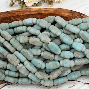 May include: A collection of light blue, barrel-shaped beads with a weathered appearance. Each bead has a slightly irregular shape and is strung together. The beads are displayed in a wooden bowl, suggesting a craft or jewelry-making theme.