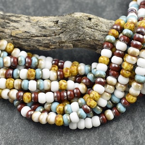 May include: A strand of colorful seed beads in white, brown, yellow, and blue. The beads are arranged in a random pattern and are strung on a clear thread.