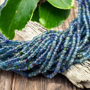 May include: A collection of small, translucent glass seed beads in various shades of blue, ranging from light sky blue to deep navy, with occasional hints of green, strung together in multiple strands to form a textured, multi-layered cluster.