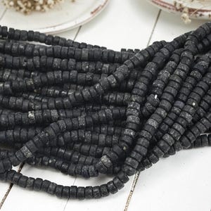 May include: A collection of multiple strands of small, cylindrical black beads with a matte, textured finish. The beads are arranged in a dense, overlapping pile, showcasing their uniform shape and dark, weathered appearance across the entire group.