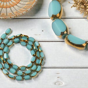 May include: A turquoise and gold beaded necklace is displayed on a white wooden surface. The necklace features oval-shaped beads with gold accents. A decorative bowl and dried flowers are also visible.