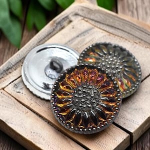 May include: Three round buttons featuring a sunflower-inspired design with dark, iridescent petals and a textured center. One button is flipped to show a smooth, reflective silver-toned back with a central shank for attachment.