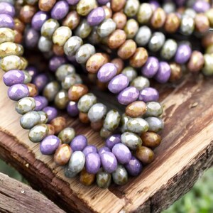 May include: A close-up of a strand of glass beads in shades of purple, green, and brown. The beads are faceted and have a slightly iridescent finish.