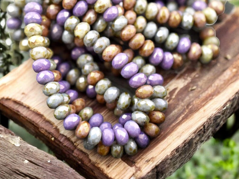 May include: A close-up of a strand of glass beads in shades of purple, green, and brown. The beads are faceted and have a slightly iridescent finish.