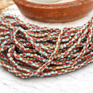 May include: Strands of colorful beads with a repeating pattern of red, light blue, and yellow stripes. The beads are small and cylindrical, and are arranged in multiple strands. A wooden bowl is in the background.