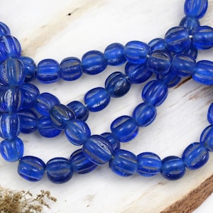 May include: Strands of vibrant blue glass beads with a ribbed design. The beads are oval-shaped and strung together, creating a necklace or bracelet. The beads are displayed on a wooden surface.