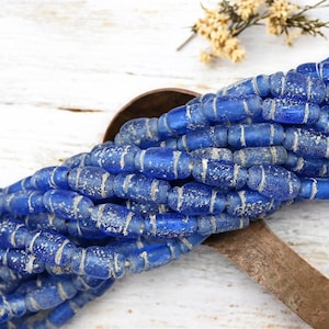 May include: Strands of blue glass beads with white stripes and speckled accents. The beads are oval-shaped and strung together, creating a textured and visually appealing design. A rustic wooden spoon is in the background.