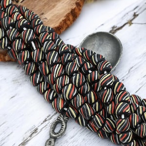 May include: Strands of black beads with red, yellow, and white stripes. The beads are arranged on a white wooden surface, with a silver spoon and a piece of wood in the background. The beads are likely for jewelry making.