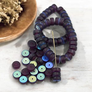 May include: An overhead shot displays a collection of dark purple, cylindrical beads with iridescent blue and green highlights. Some beads are strung, others scattered. A gold needle is inserted through a stack of beads, suggesting jewelry making. A wooden bowl with dried flowers is in the background.