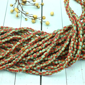 May include: Strands of colorful beads in shades of red, yellow, and light green. The beads are cylindrical with a striped pattern. The beads are arranged in a pile on a white wooden surface. A sprig of yellow berries is in the background.