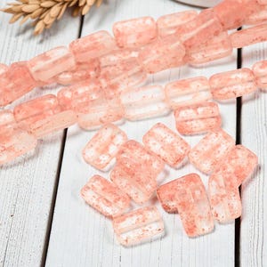 May include: Strands of rectangular, translucent pink beads with a speckled appearance. The beads are strung together, with some loose beads scattered nearby. The beads have a slightly frosted look, and are likely for jewelry making.