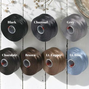 May include: Spools of thread in various colors, including black, charcoal, grey, chocolate, brown, light copper, and light blue. Each spool is labeled with its color name. The thread is tightly wound on a cylindrical core.
