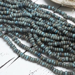 May include: A collection of multiple strands of small, round, disc-shaped beads in varying shades of blue, gray, and teal. Each bead features a textured, speckled surface with a matte finish, arranged in long, parallel lines across the frame.