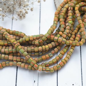 May include: A long strand of small, cylindrical glass beads featuring a multi-colored striped pattern in shades of olive green, orange, yellow, and blue. The beads are arranged in a loose, coiled formation, showcasing their repeating vertical color bands.