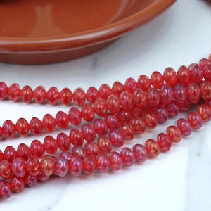 May include: Strands of red, oval-shaped beads with a glossy finish. The beads are strung together, creating a flowing pattern. A terracotta-colored bowl is visible in the background. The beads are suitable for jewelry making.