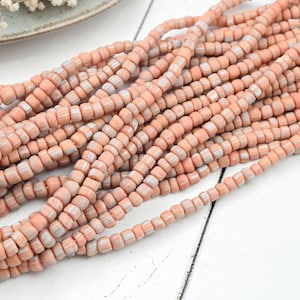 May include: A collection of peach-colored beads strung together on multiple threads. Each individual bead features a cylindrical shape with a matte finish and subtle vertical ridges, displaying a consistent light orange hue throughout the strands.