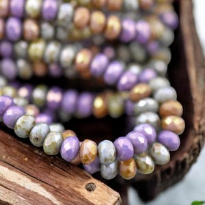 May include: A close-up of a strand of beads in shades of purple, gold, and gray. The beads are small and have a faceted, iridescent finish.