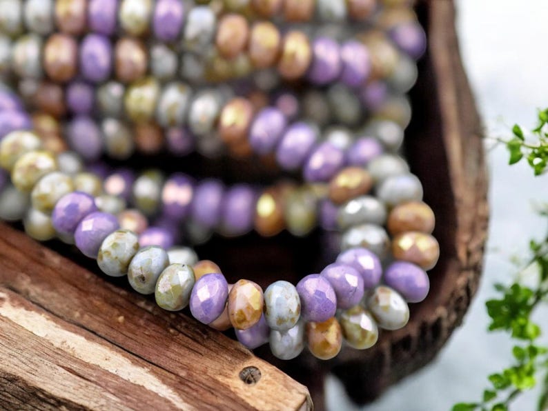 May include: A close-up of a strand of beads in shades of purple, gold, and gray. The beads are small and have a faceted, iridescent finish.