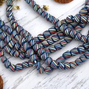 May include: Strands of blue, red, yellow, and white striped beads. The beads are oval-shaped and strung together, creating a textured and colorful pattern. The beads are arranged on a white wooden surface.