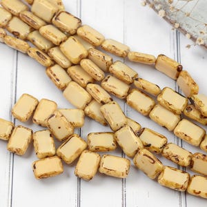 May include: Cream-colored rectangular beads with brown accents are scattered across a white wooden surface. The beads are strung together in strands, suggesting they are for jewelry making. The beads have a slightly aged, vintage appearance.