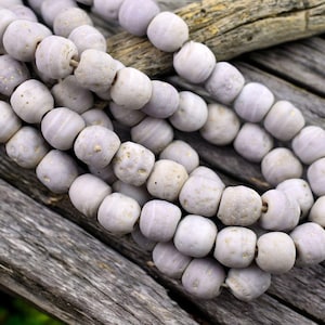 May include: A strand of light purple glass beads with a textured surface. The beads are round and have a slightly irregular shape.