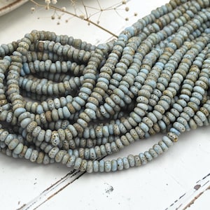May include: A collection of multiple strands of small, round, disc-shaped beads. The beads feature a light blue color with a textured, speckled finish in shades of brown and tan, giving them a porous, stone-like appearance arranged in loose piles.